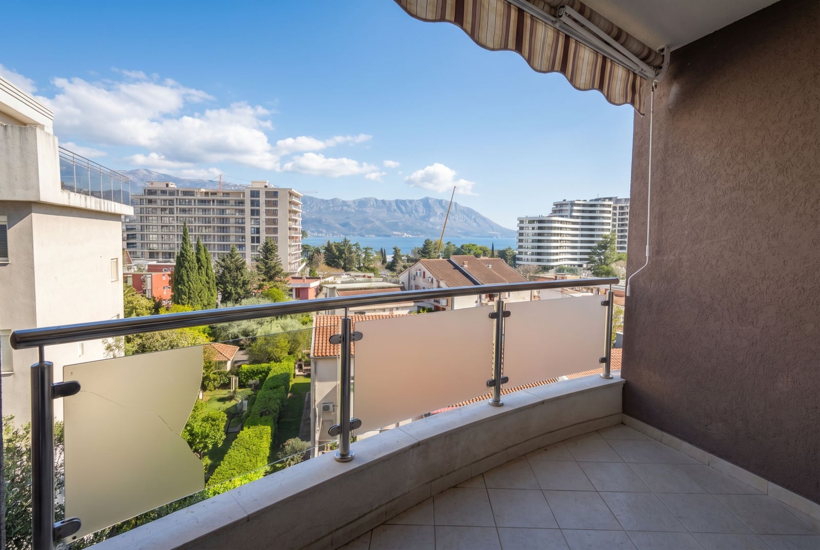 Apartment balcony in Budva, Montenegro, with panoramic views of the sea, mountains, and city, protective canopy, and glass railing.