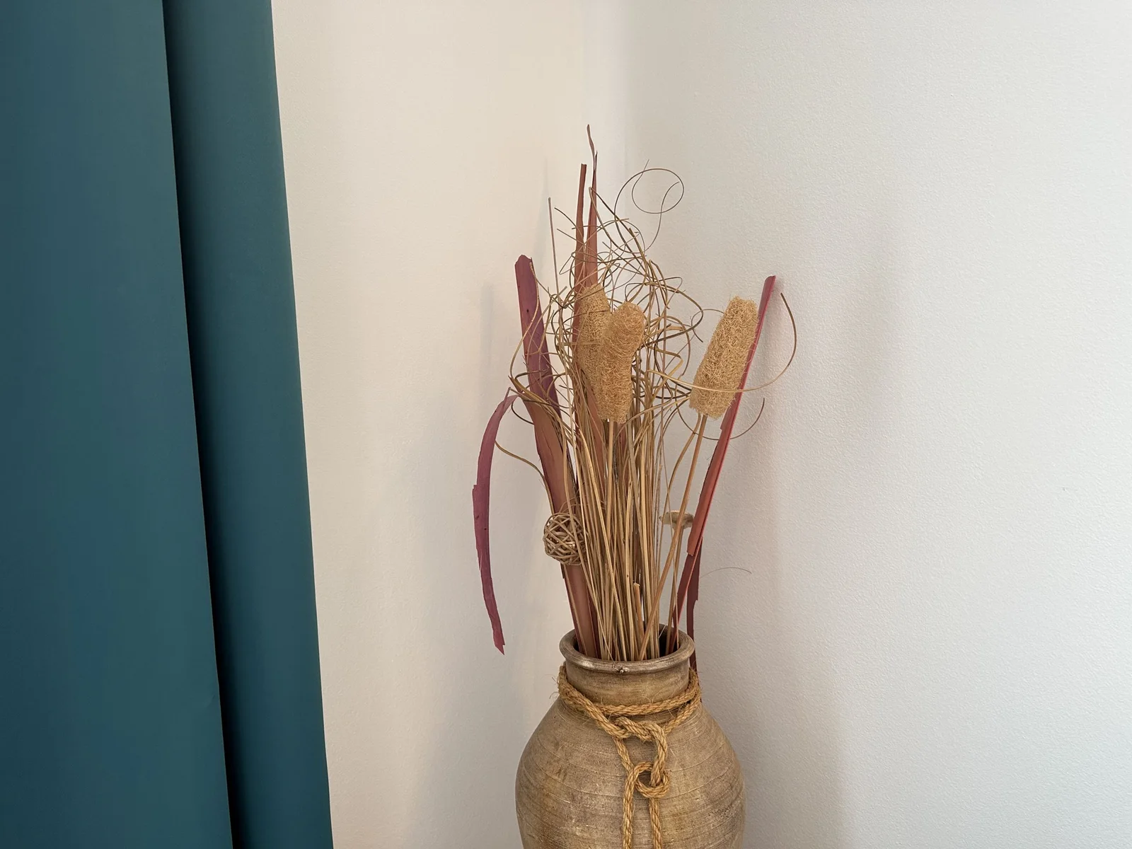 Decorative vase with dried plants against a wall. Part of a blue curtain and white surface visible.