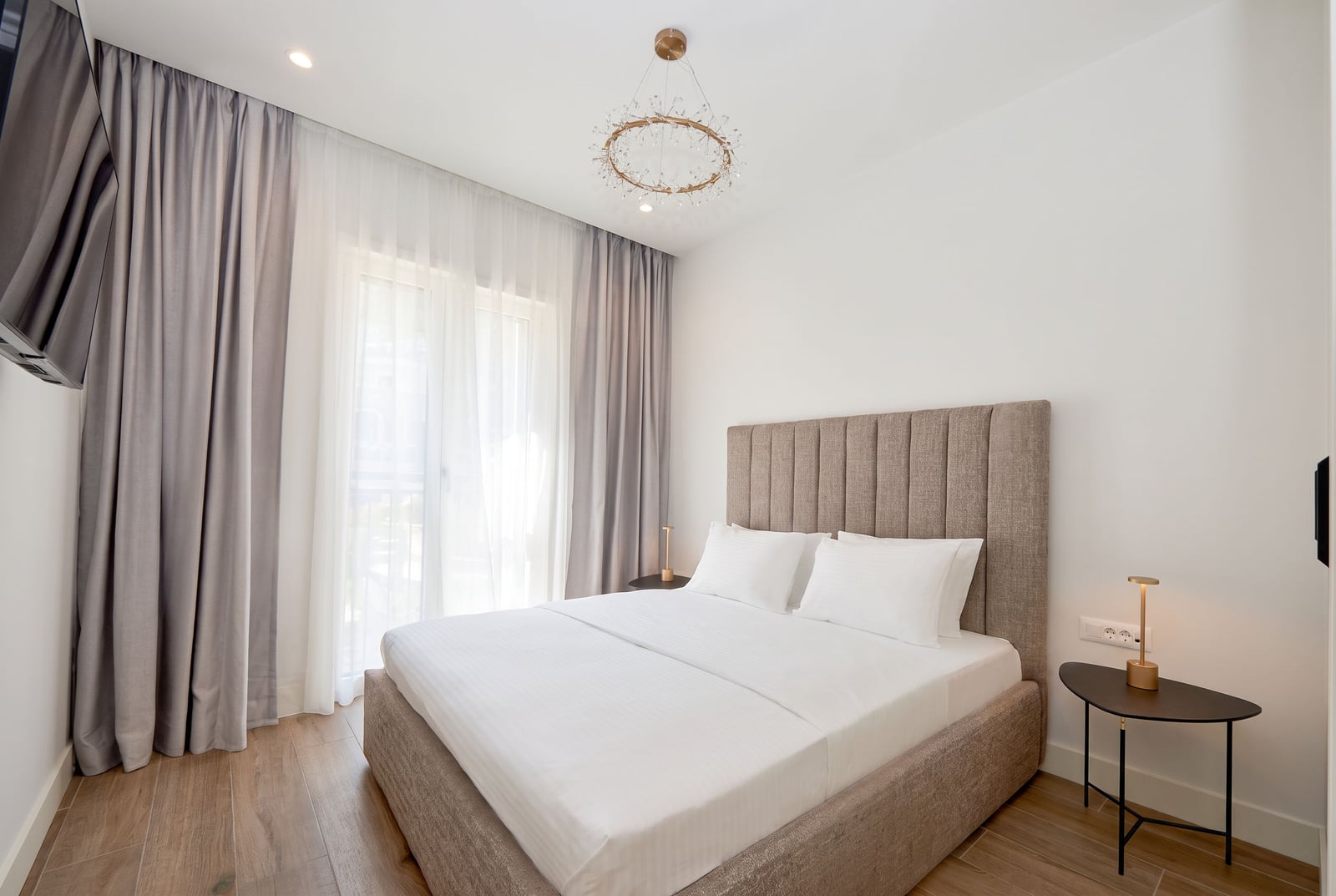 Modern bedroom featuring a double bed with upholstered headboard, circular chandelier, and floor-length curtains.