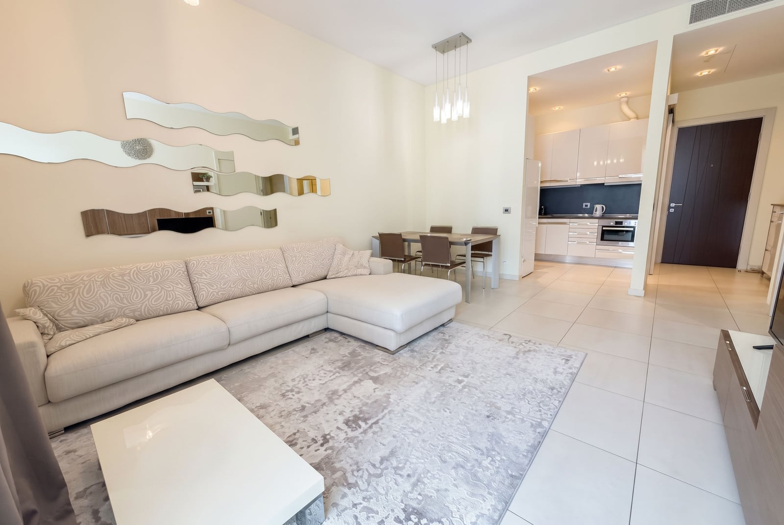Bright living room with a large beige sectional sofa, patterned rug, wavy wall mirrors, and open kitchen view.