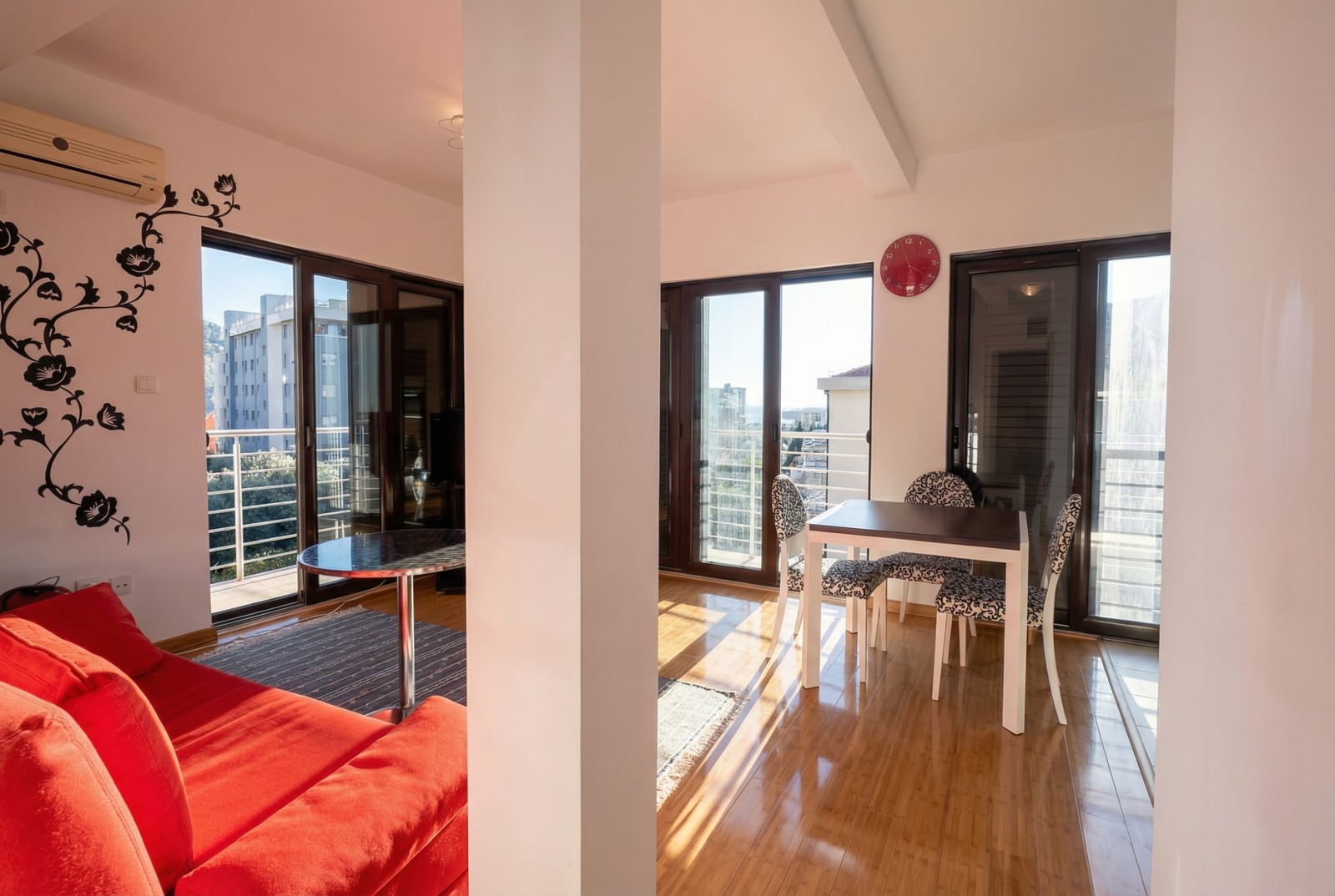 Bright living room with a red sofa and dining area in an apartment in Bečići, Montenegro, with access to the balcony.