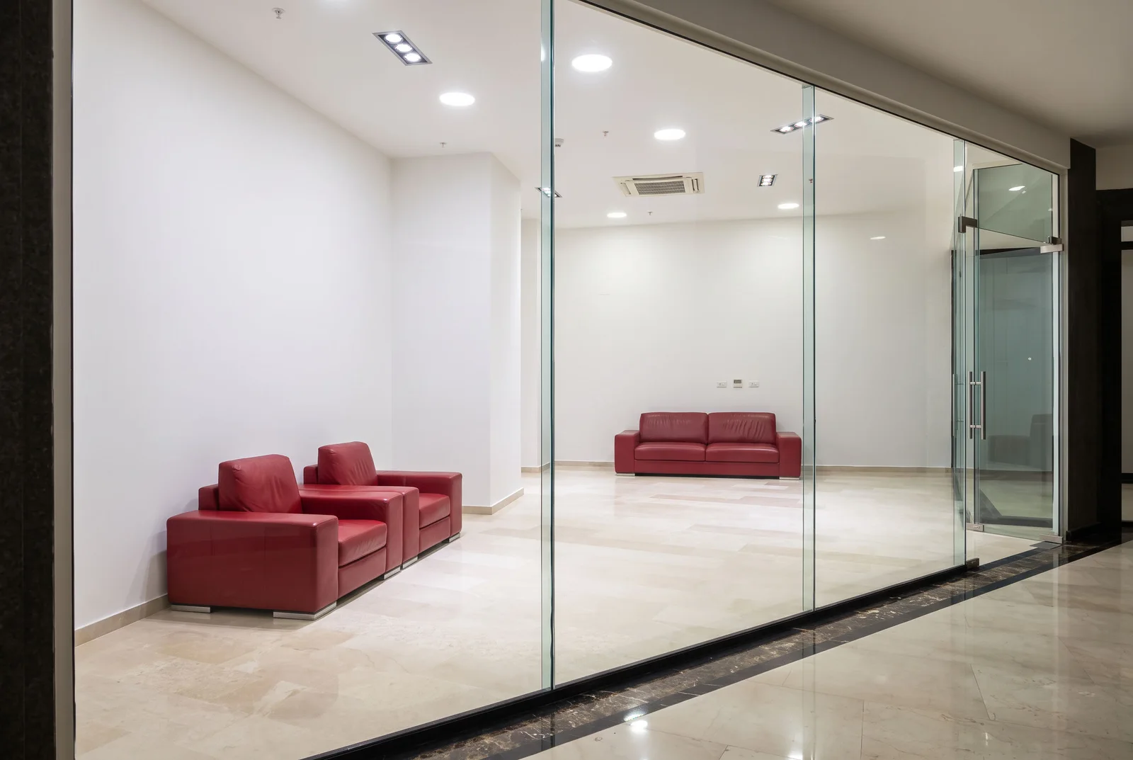 Office space with red leather chairs and sofa behind glass partitions and marble flooring.