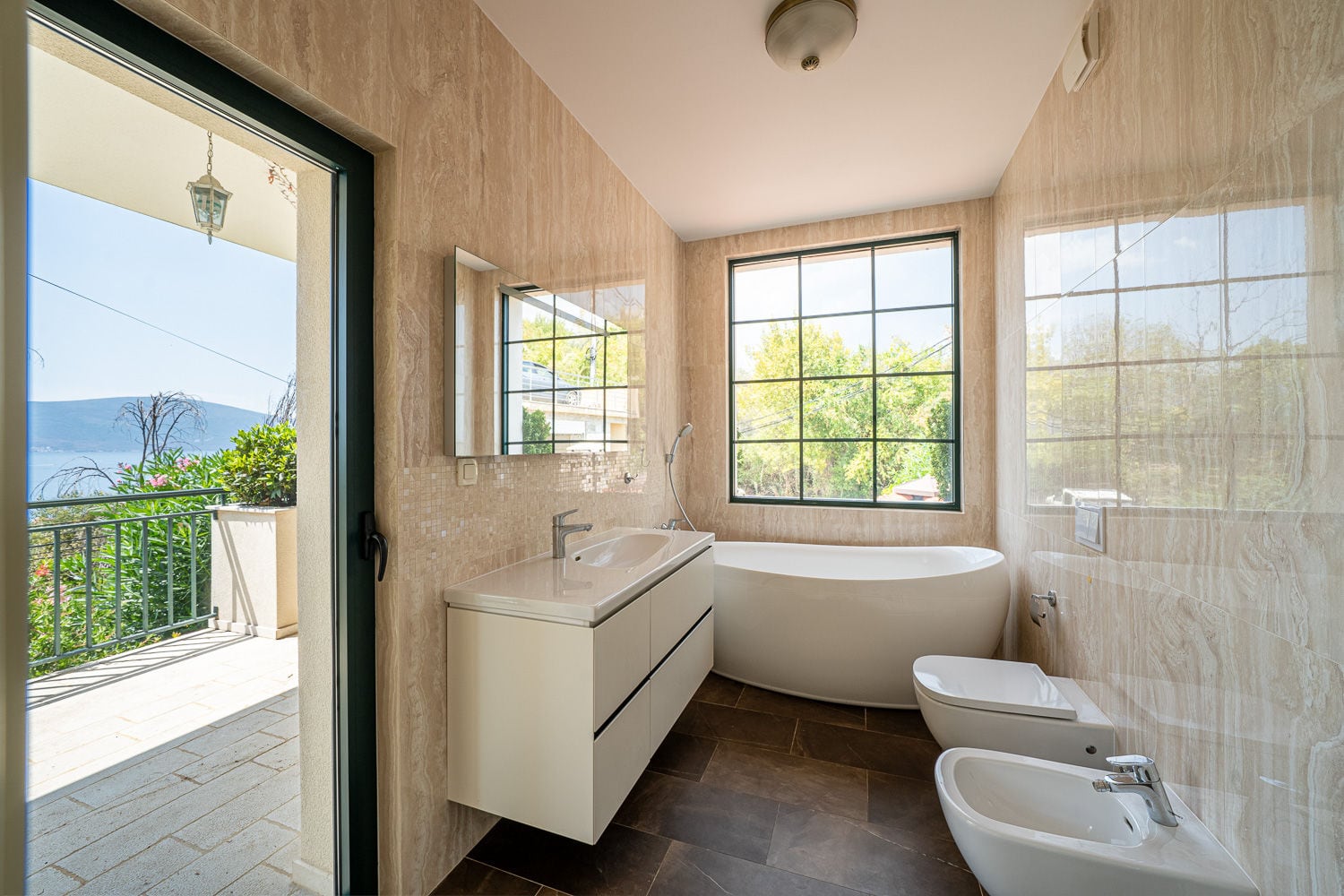 Modern bathroom featuring a freestanding tub, vanity sink, toilet, and bidet. An open door reveals a balcony with a scenic water view.