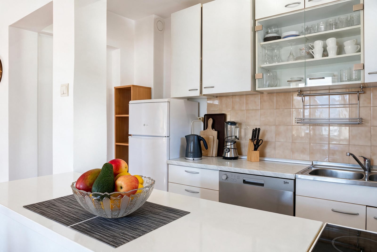 Modern kitchen with white cabinets, fridge, dishwasher, sink, and a fruit bowl on the central island.