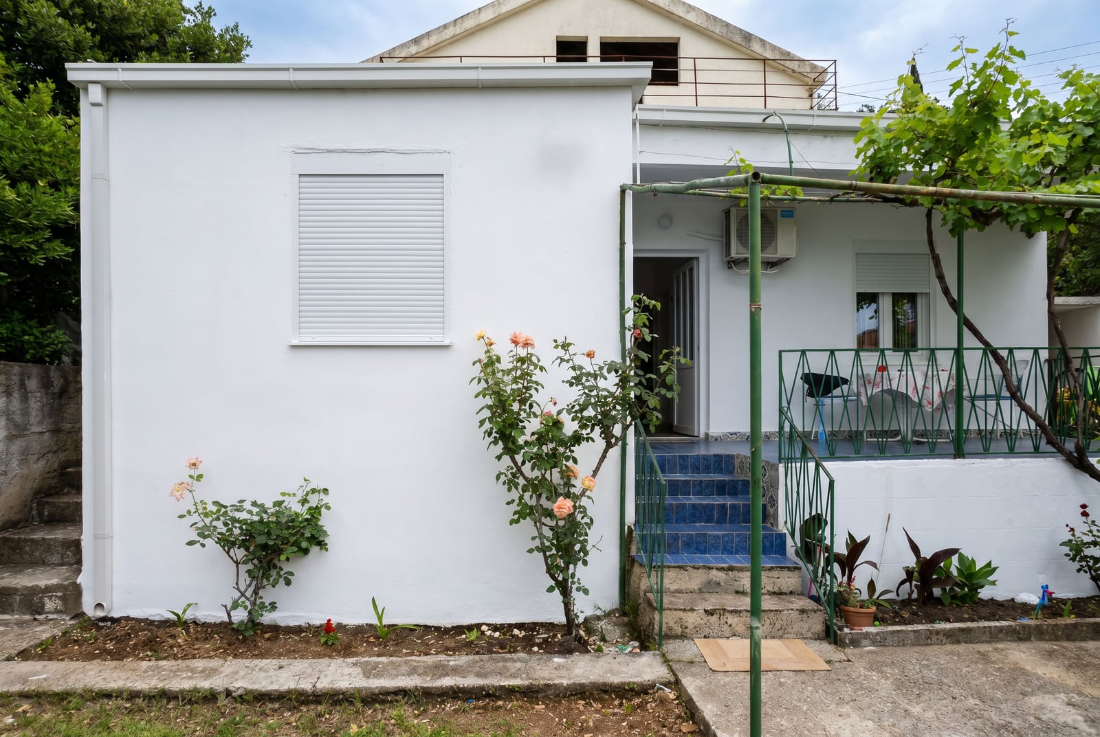 White house with a terrace, blue tiles, and a garden in Sutomore, Montenegro. Facade with roses and an outdoor seating area.