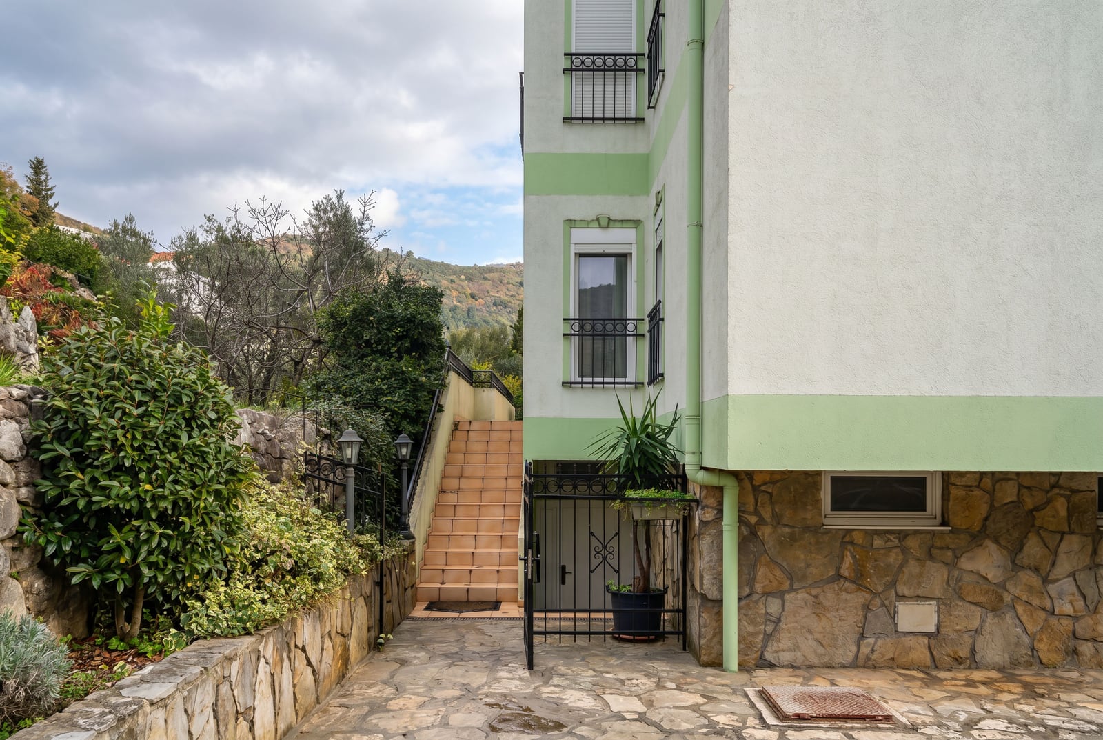 Facade of a house in Budva, Montenegro: stone-finished base, exterior staircase, and apartment entrance with a green garden.