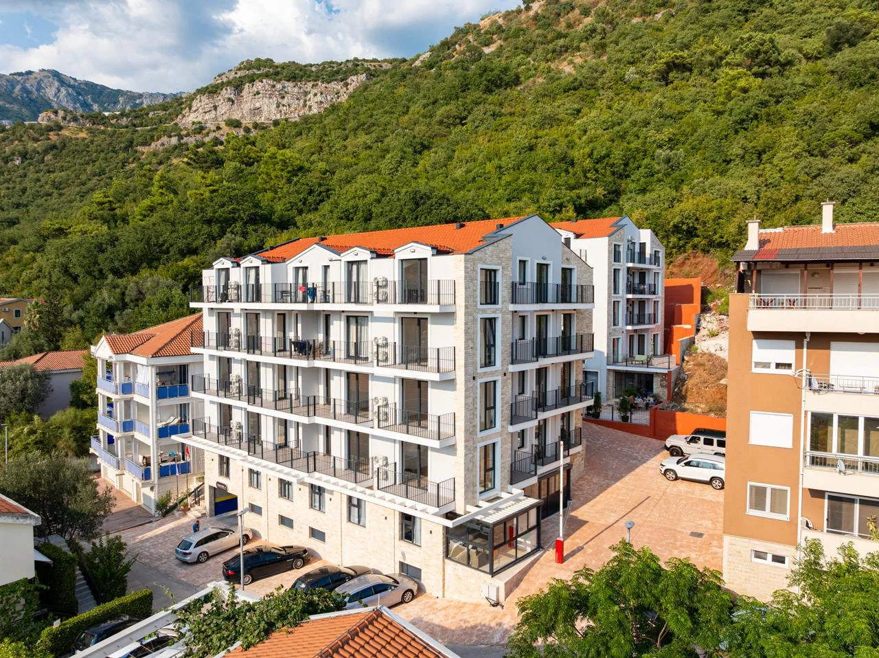 Exterior view of a multi-story apartment building with balconies and parking against a green hillside.