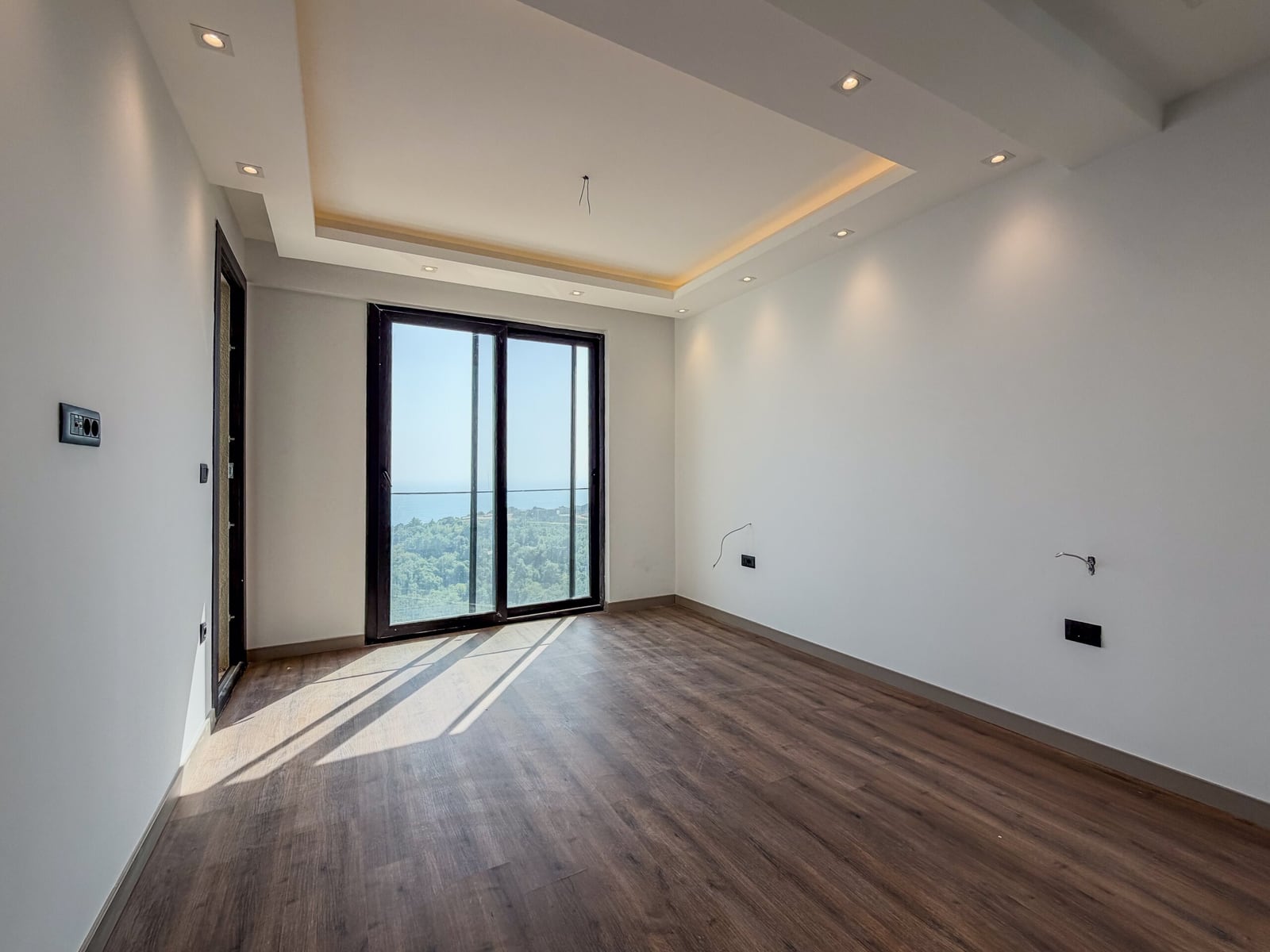 Empty room with wood flooring, white walls, recessed lighting, and a large window overlooking the sea.