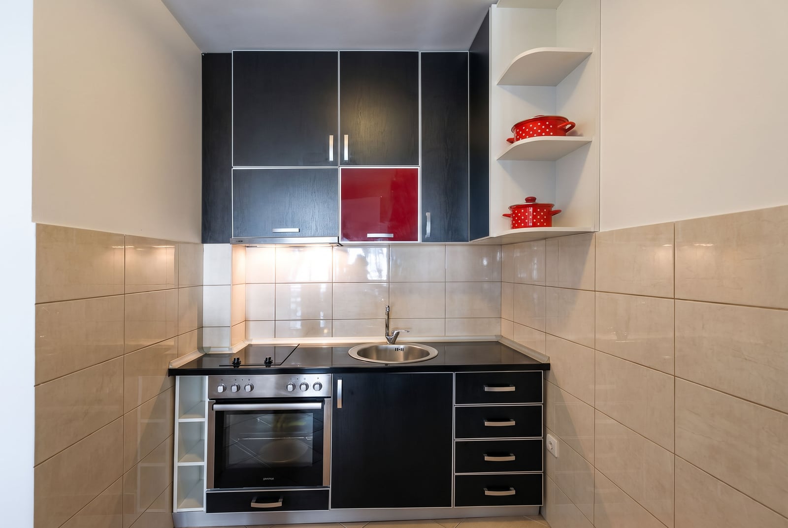 Compact kitchen with black cabinets, red accent panel, stove, oven, sink, and beige tiled walls.