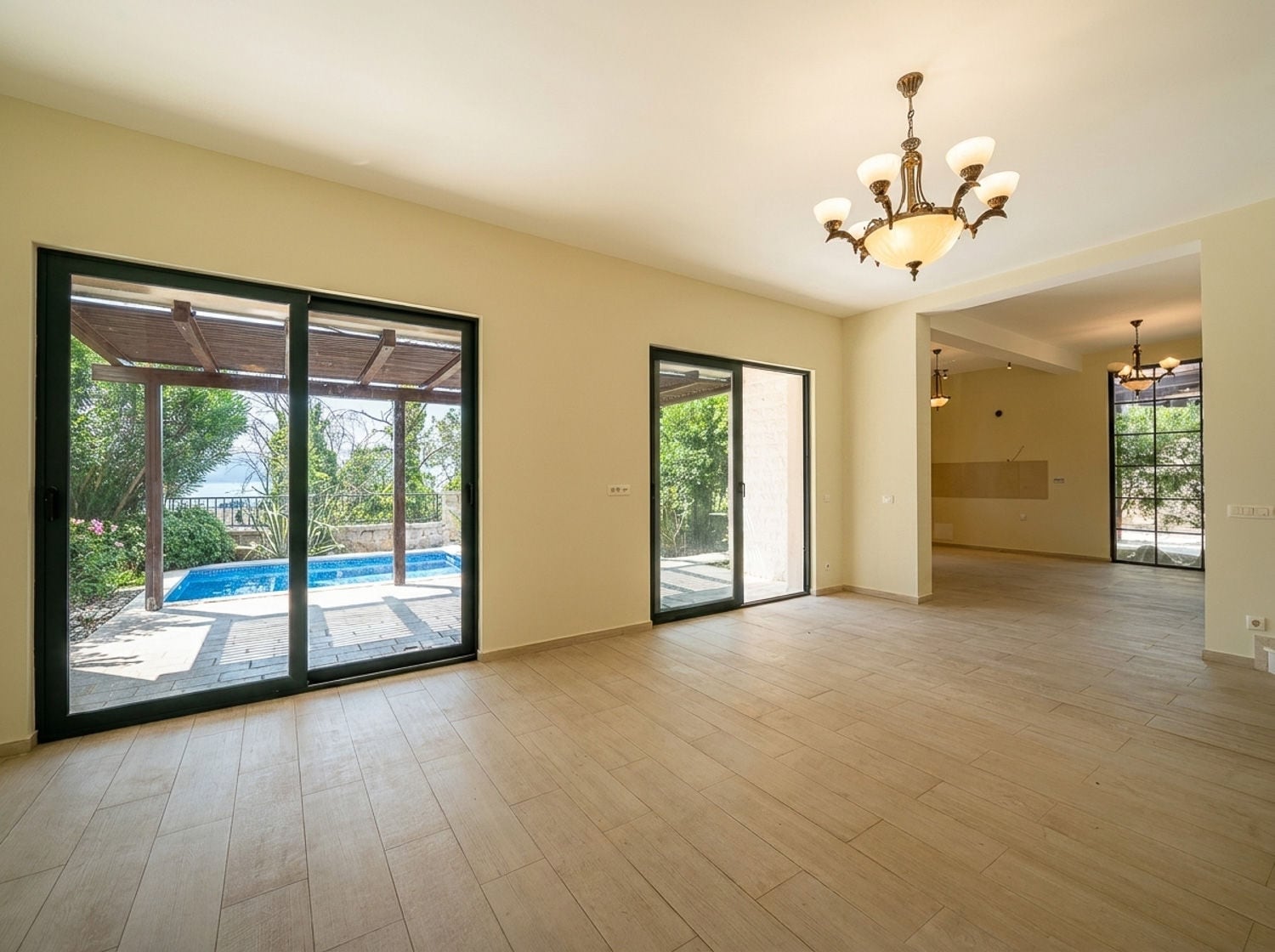 Empty room with wood-look tile floor and chandelier, large sliding doors open to a pool terrace.
