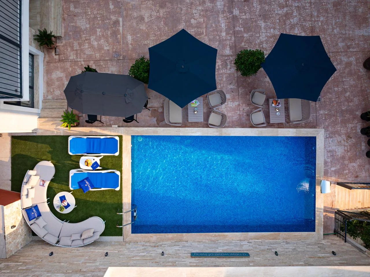 Top-down view of a blue swimming pool surrounded by a patio with umbrellas, sunbeds, and curved outdoor furniture on grass.