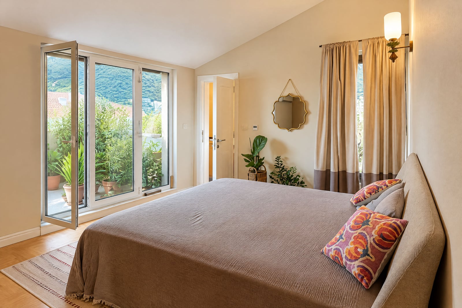 Bright bedroom featuring a large bed, decorative mirror, and an open door leading to a balcony with plants.