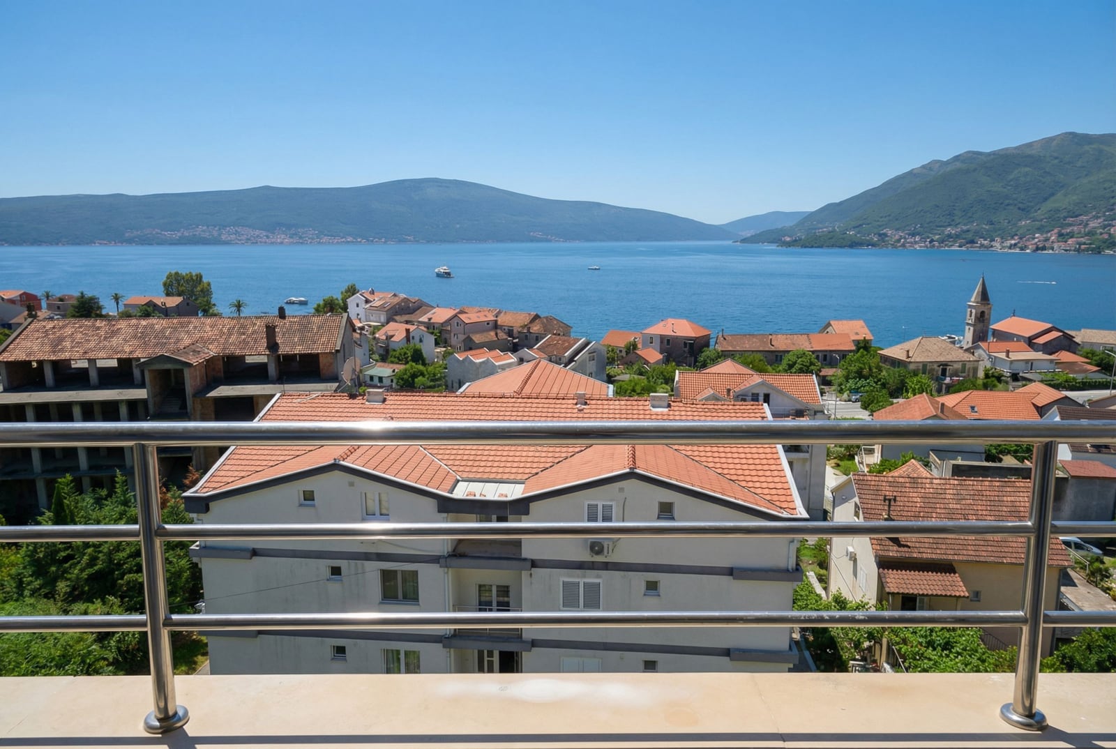 View from the apartment balcony in Tivat, Montenegro: panoramic view of the Bay of Kotor, terracotta roofs, and mountains.