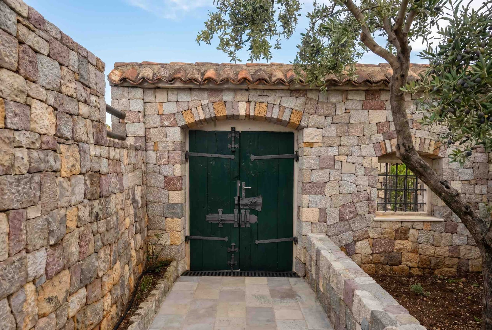 Entrance to a stone house in Luštica, Montenegro: massive green door, Mediterranean stonework, and an olive tree.