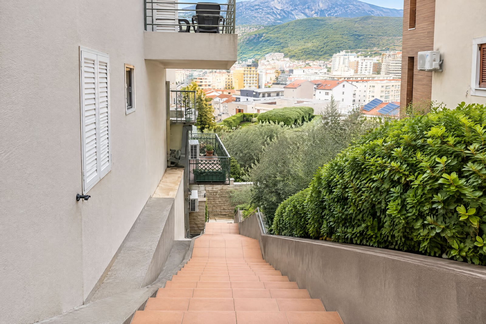 Exterior view showing a terracotta tiled staircase, white shutters, balconies, and a cityscape with mountains in the background.