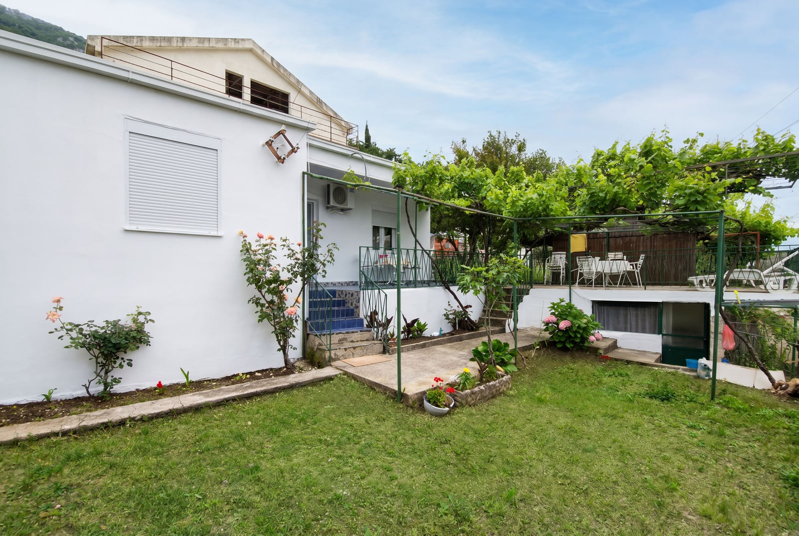 White house with a terrace, garden, and grapevine pergola in Sutomore, Montenegro. Cozy courtyard with a lounge area and flowers.