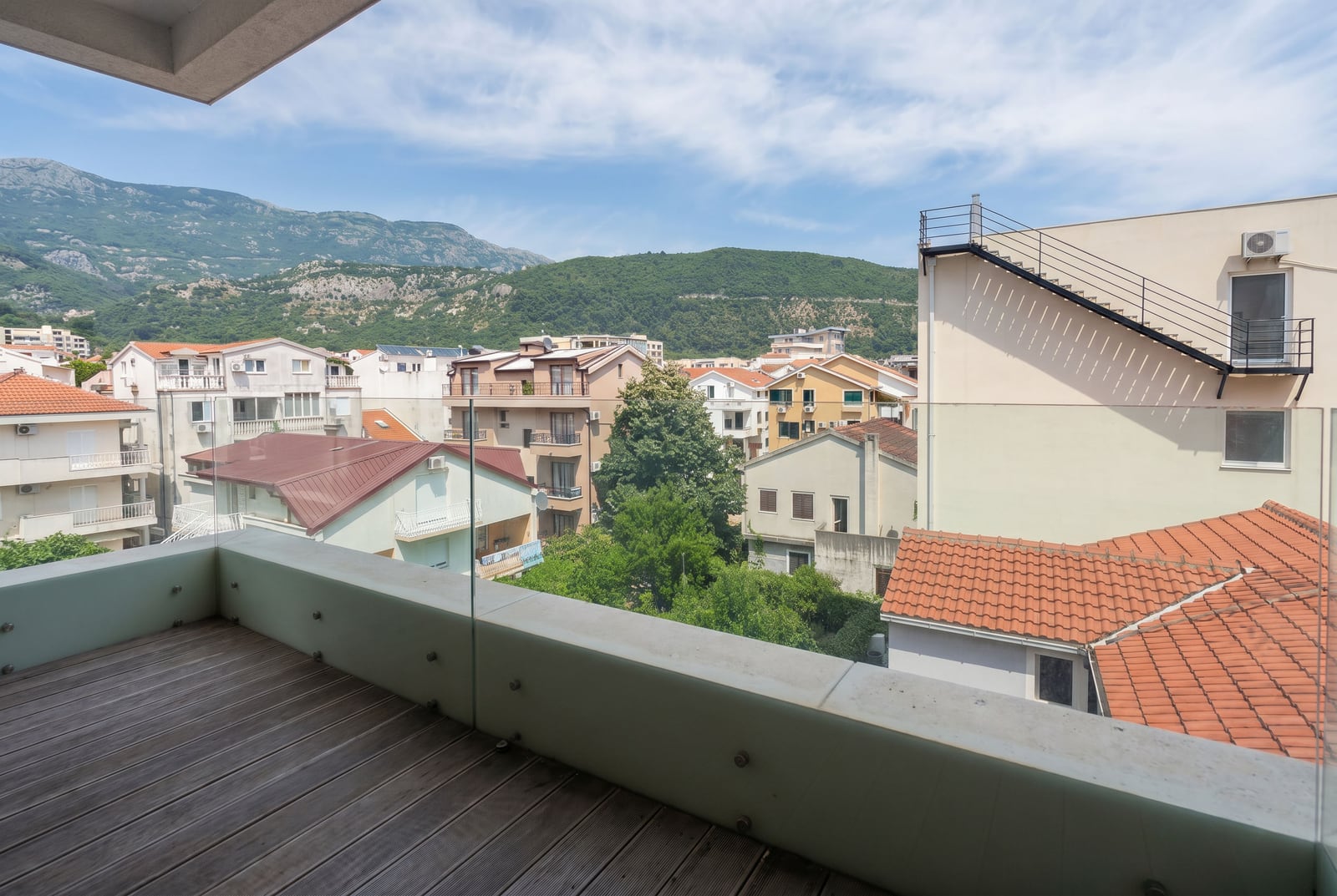 Balcony with wooden decking and glass railing in a Budva apartment, Montenegro, featuring mountain and city views.