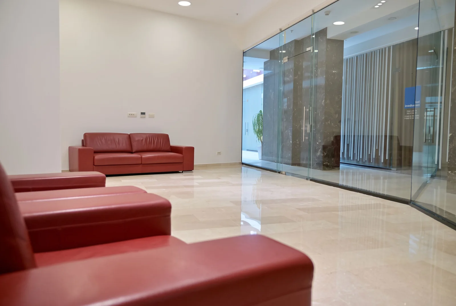 Modern lobby or waiting area featuring red leather sofas, polished marble floors, and large glass partition walls.