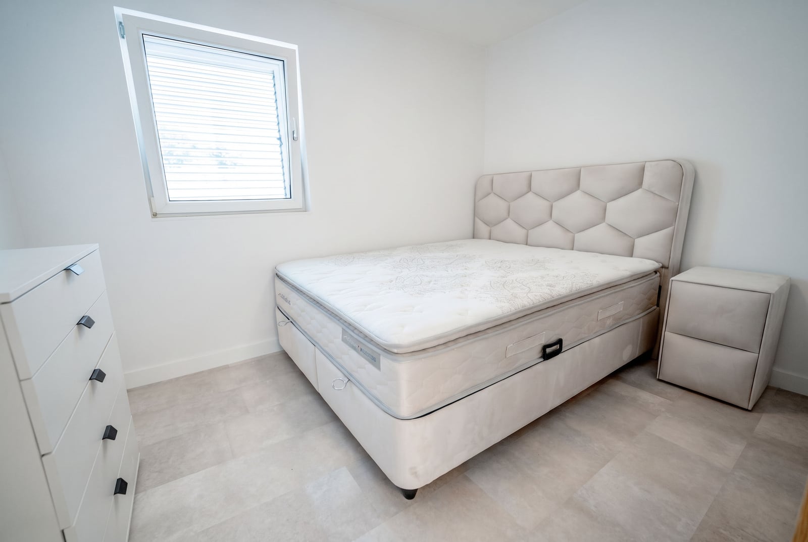 Bright bedroom in an apartment, Sveti Stefan, Montenegro: double bed with upholstered headboard, dresser, and window with blinds.