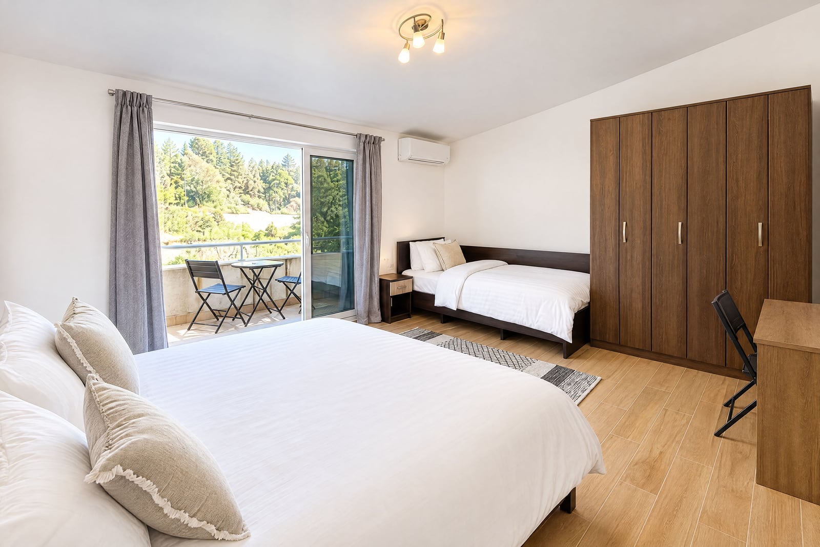 Bright bedroom featuring a double bed, single bed, large wooden wardrobe, and sliding glass door leading to a balcony with forest views.