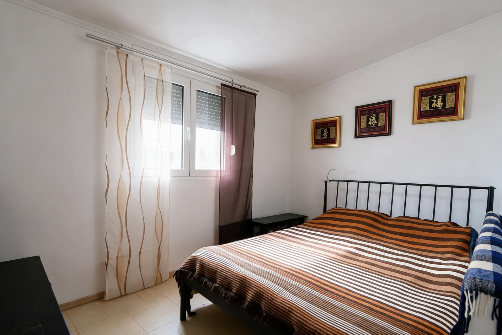 Bedroom featuring a metal bed frame with striped bedding, a window with sheer curtains, and three framed artworks on the wall.