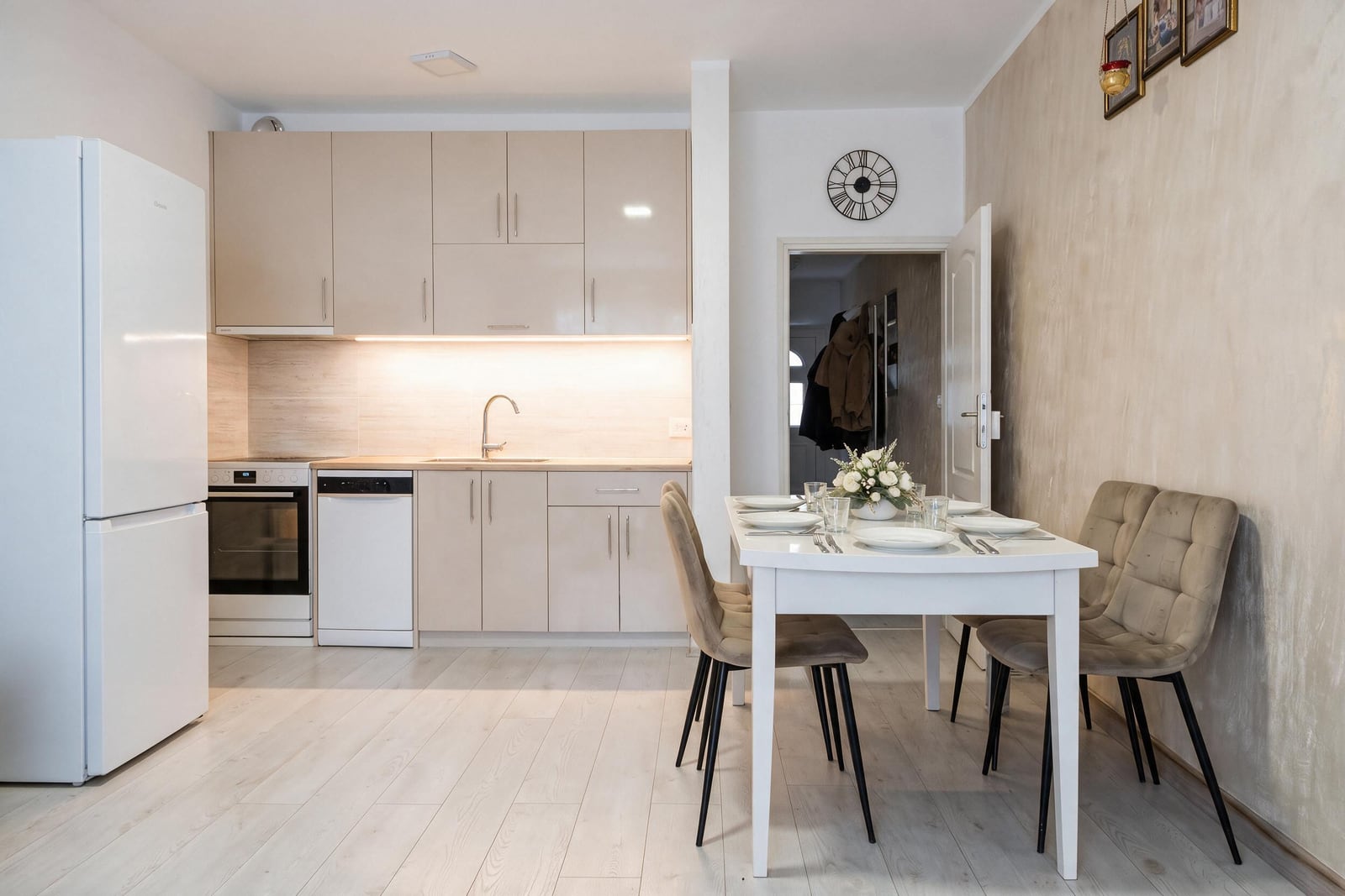 Modern kitchen with light cabinets, fridge, oven, and a dining area featuring a white table and beige chairs.