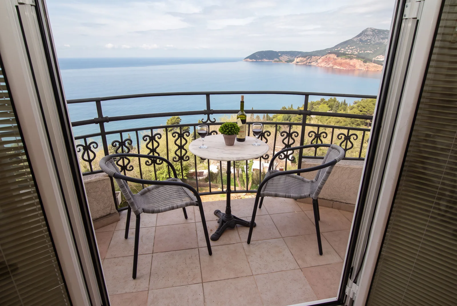 Balcony with sea view featuring a small table with wine and two woven chairs behind open doors.