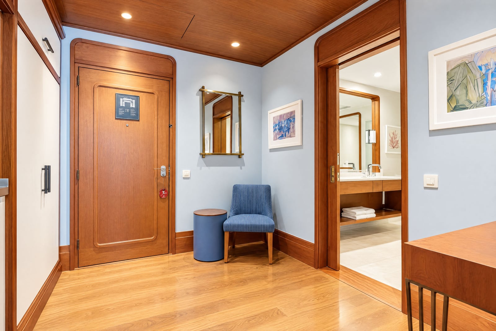 Entryway with wooden door, blue chair, and mirror; bathroom visible through doorway on the right.