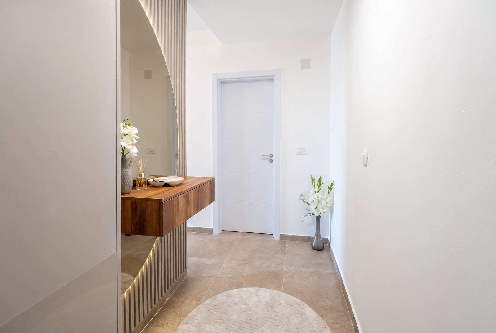 Modern hallway with a floating wooden console, round mirror, white walls, and tiled floor.