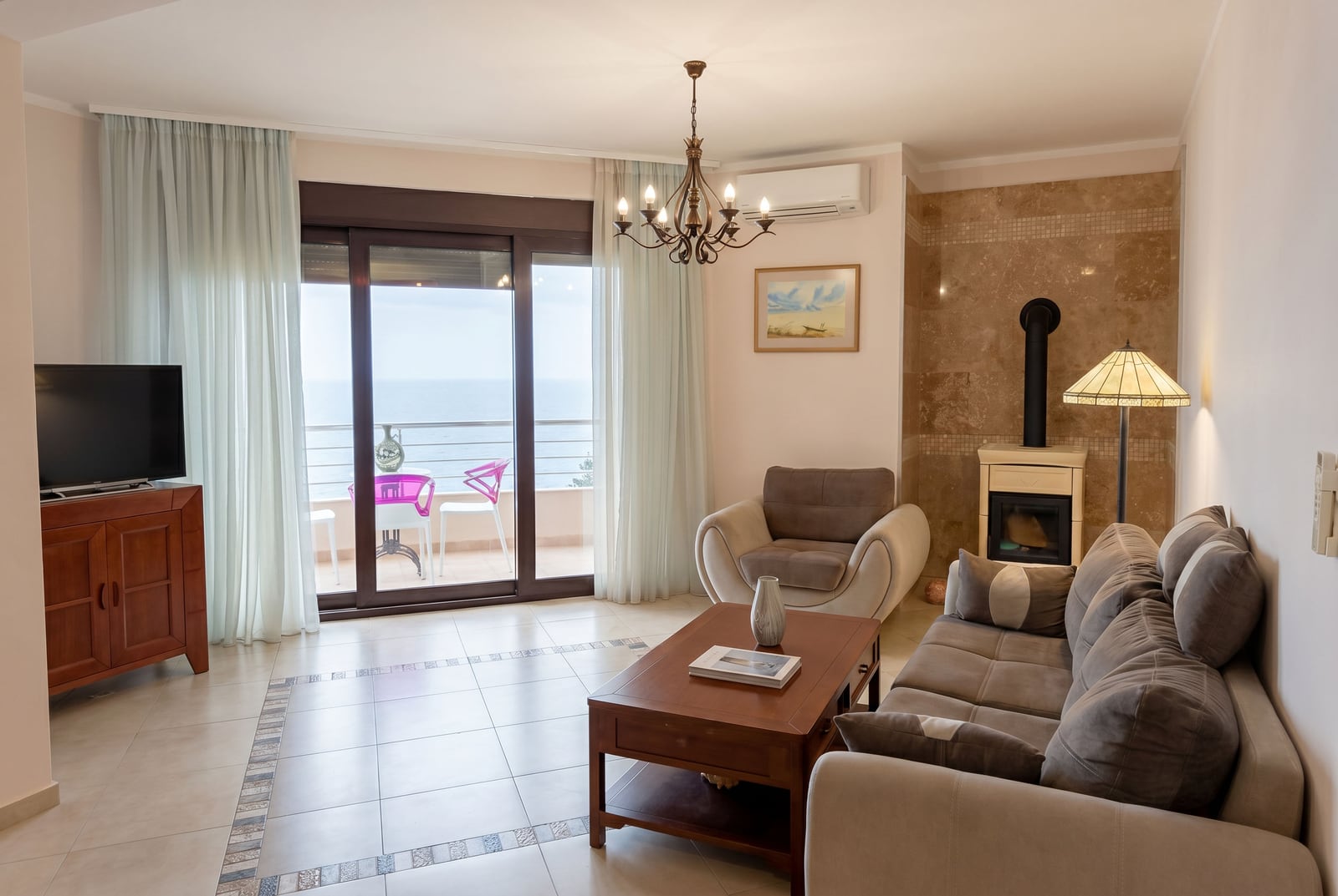 Living room in an apartment in Bar, Montenegro: upholstered furniture, fireplace, tiled floor, and access to a terrace with a sea view.