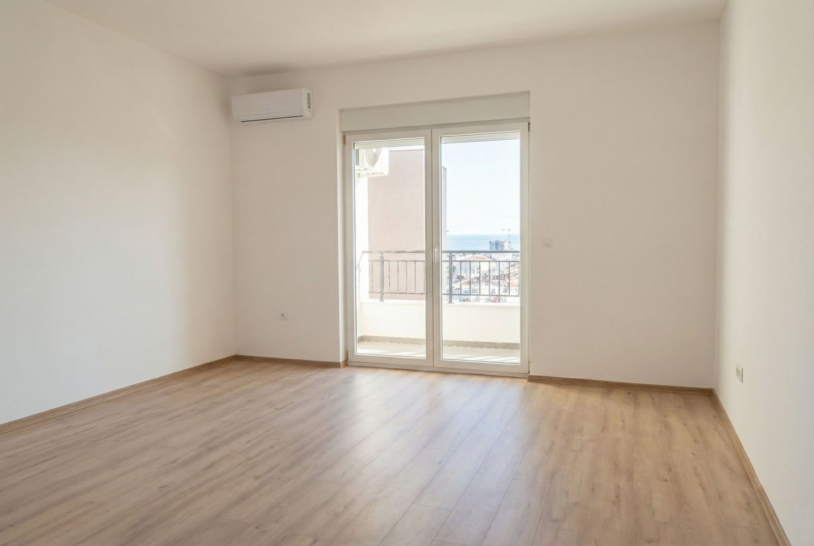 Bright empty living room with laminate flooring, air conditioning, and access to a balcony with sea views in Budva, Montenegro.