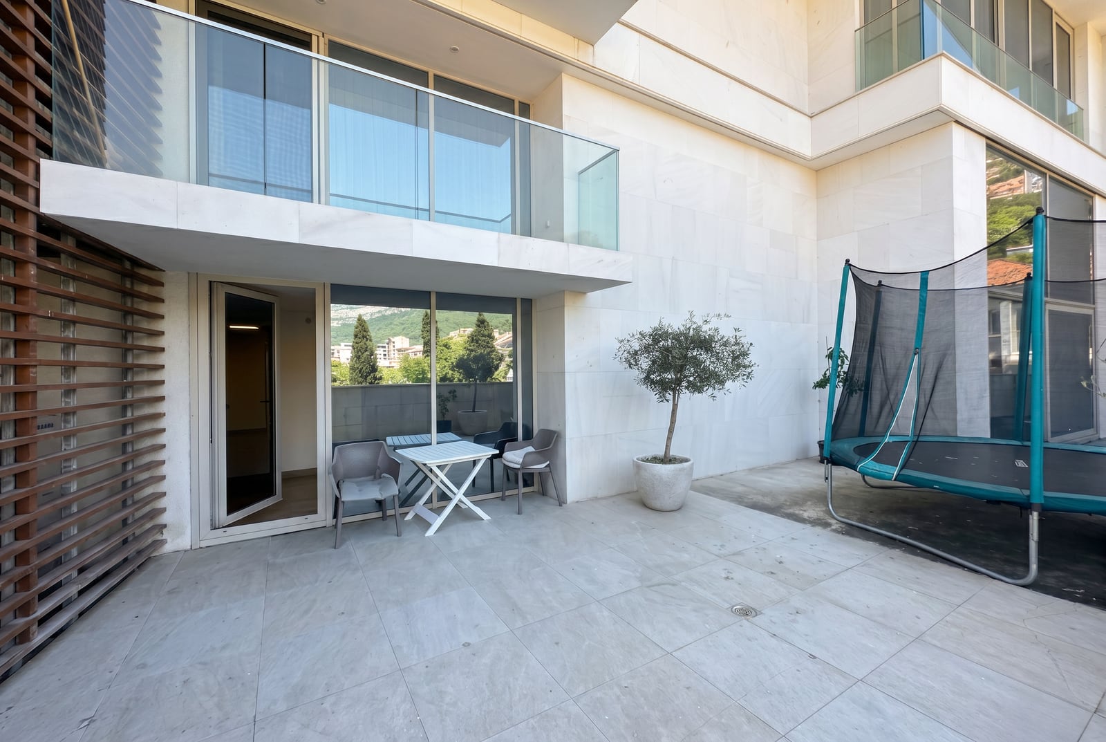 Terrace of a modern apartment in Budva, Montenegro: panoramic windows, patio furniture, and a trampoline on a stone-paved surface.
