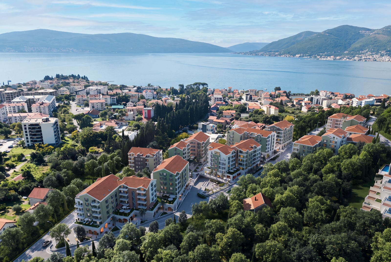 Aerial view of a coastal town featuring multi-story buildings, lush greenery, and a sea backdrop with mountains.