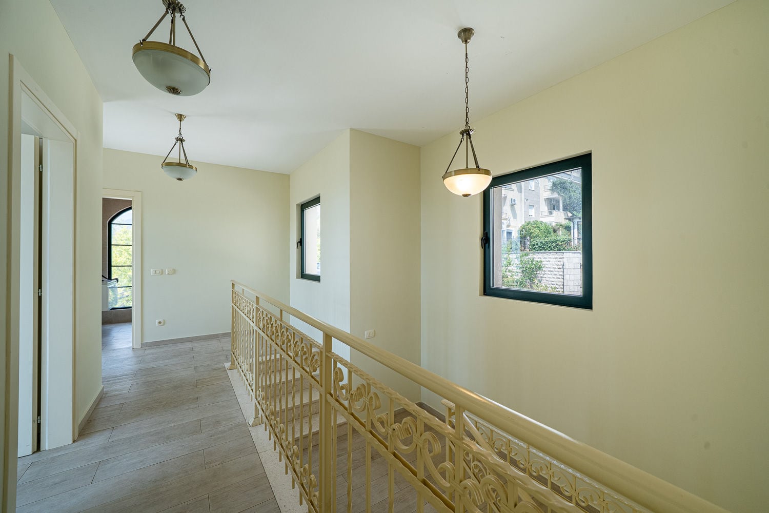 Interior hallway with staircase and ornate railing, pendant lights, windows overlooking outside.