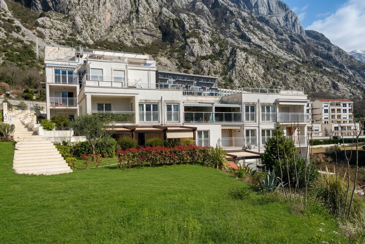 Exterior view of a multi-story residential building with balconies, situated on a green lawn at the base of rocky mountains.