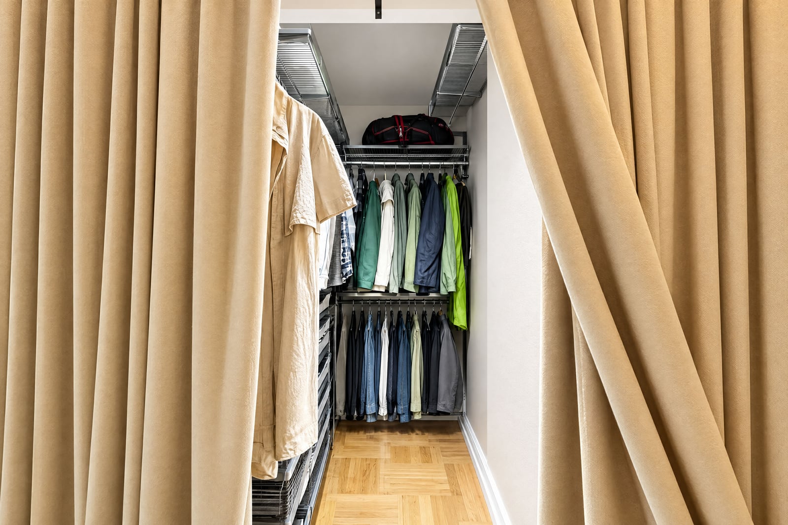 Walk-in closet with metal racks, hanging clothes, beige curtains, and a wooden parquet floor visible in the narrow space.
