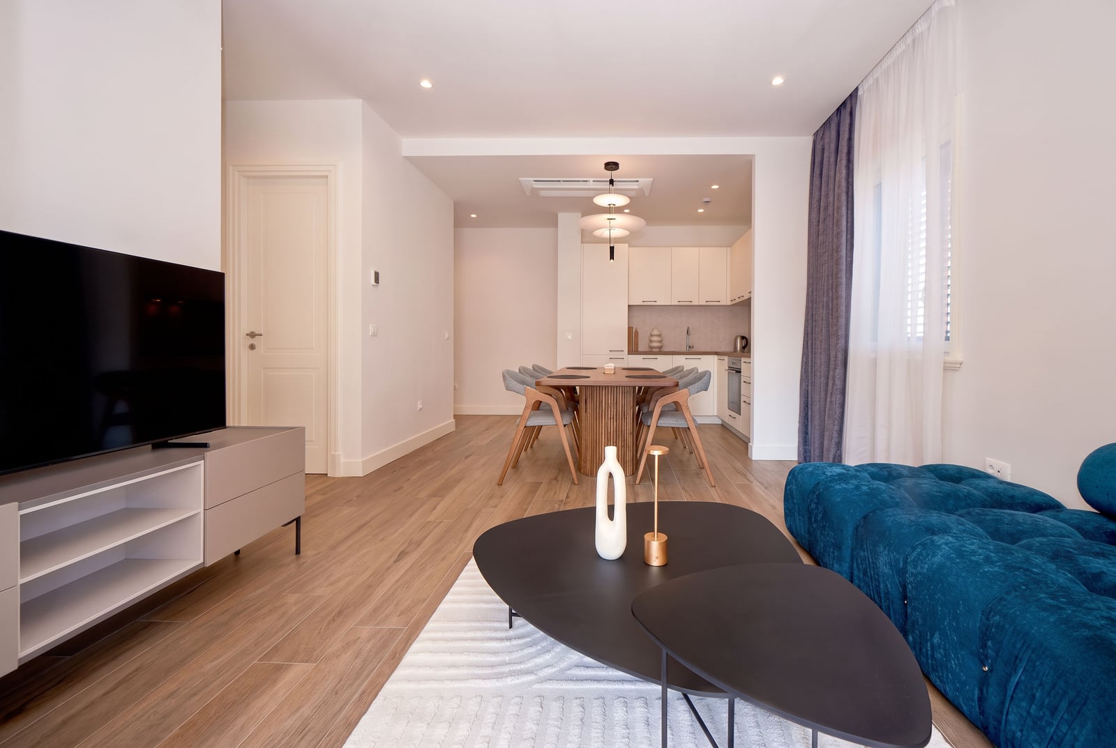 Modern living room with a blue sofa, black coffee table, and TV; dining area and kitchen visible in the background.