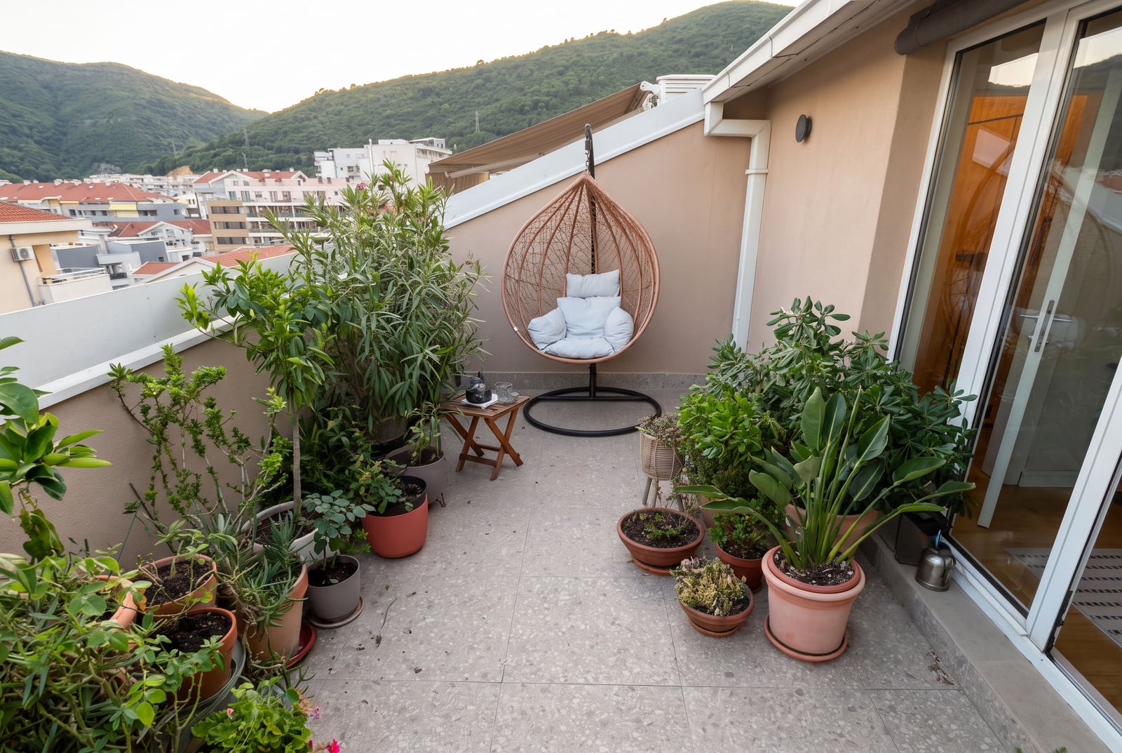 Cozy balcony featuring a hanging egg chair and numerous potted plants, overlooking residential buildings and green hills.