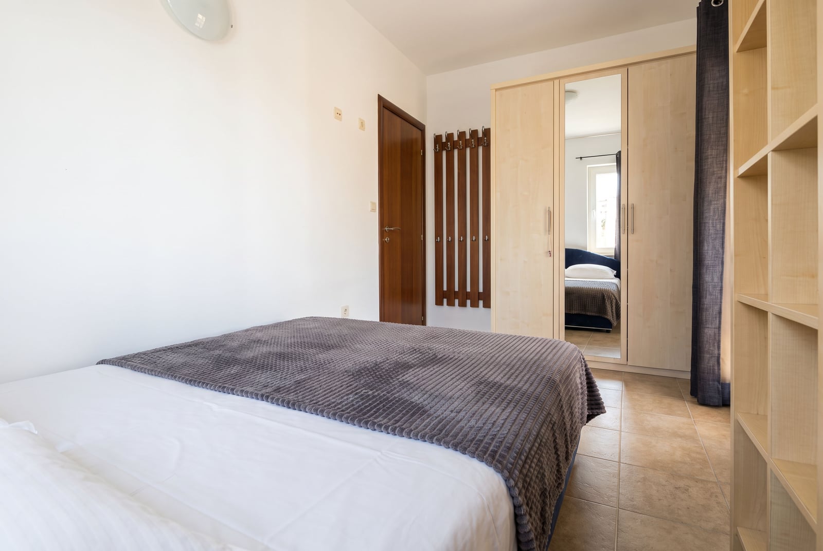 Bedroom featuring a double bed with grey throw, light wood wardrobe with mirror and open shelving.