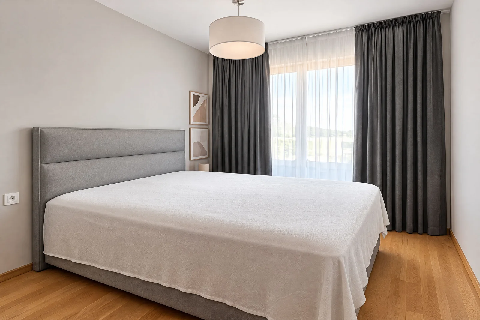 Bedroom featuring a large bed with a grey upholstered headboard, white coverlet, wooden flooring, and grey curtains by the window.
