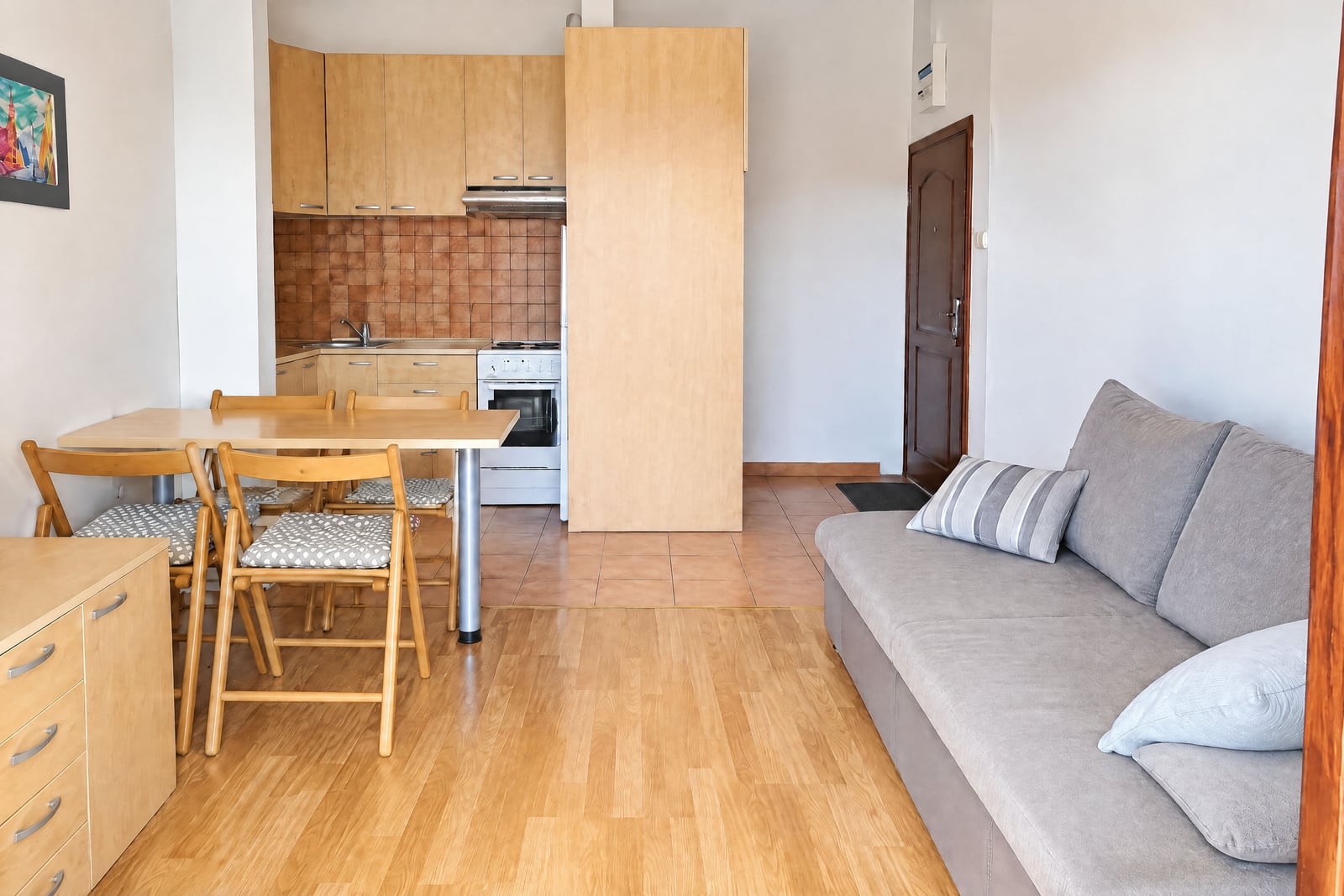 Living room with grey sofa and wooden dining table; kitchen visible in background with cabinets.