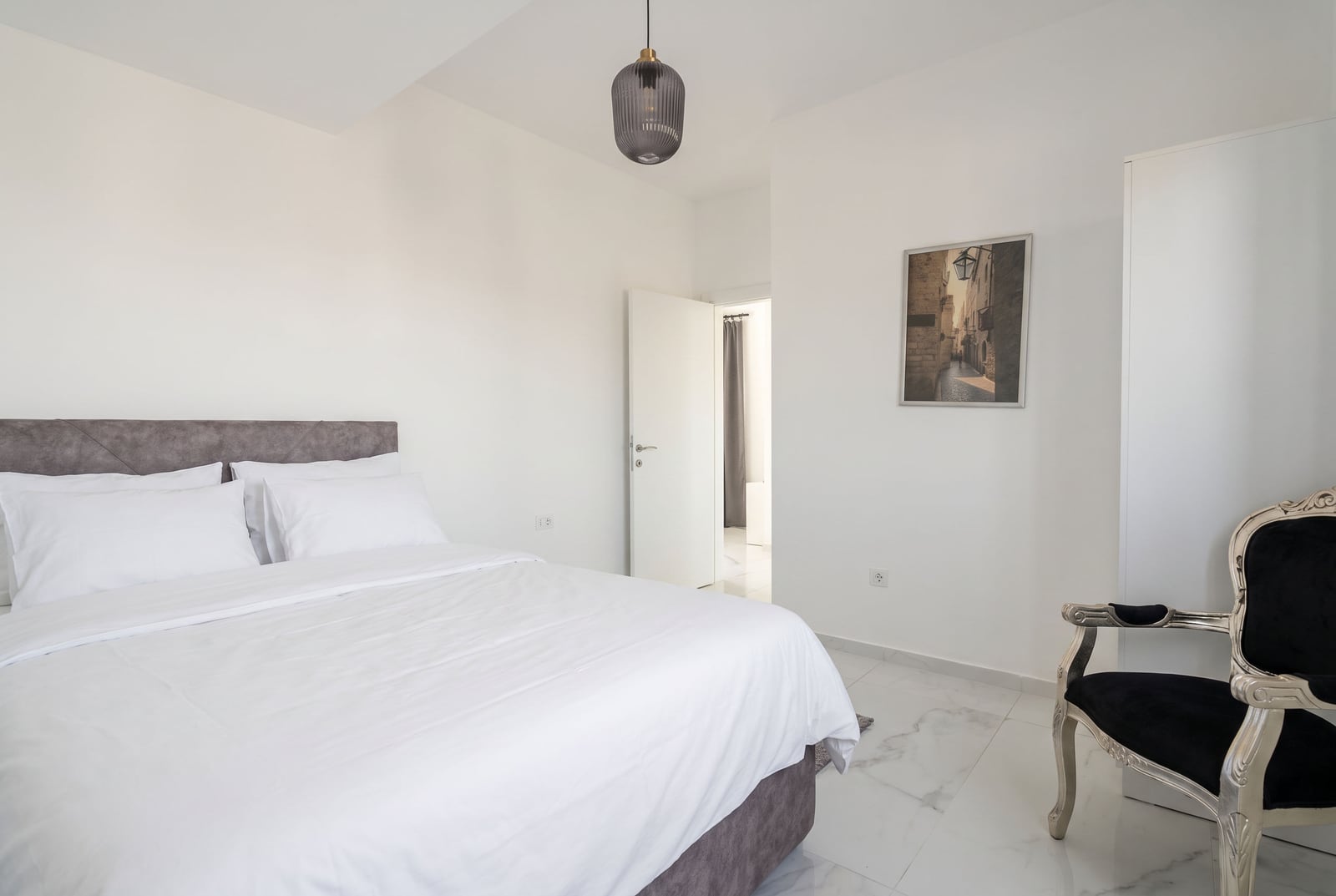Bedroom featuring a large bed with white linens, grey headboard, pendant light, and black ornate chair on marble flooring.