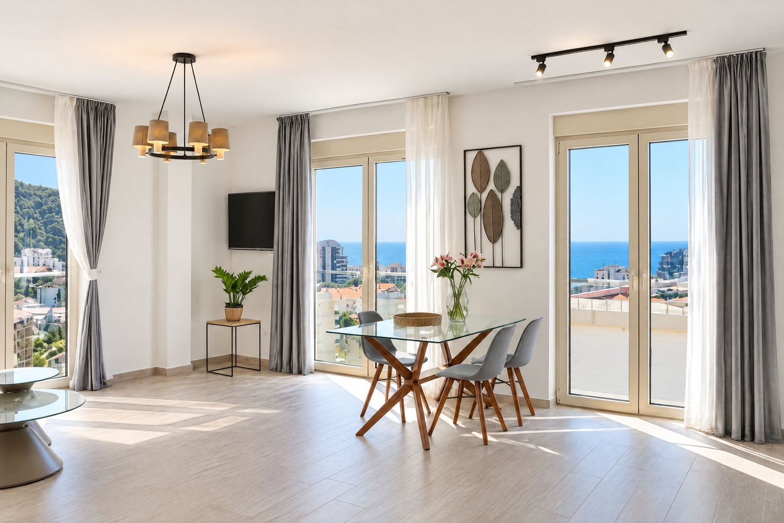 Bright room with glass dining table, chairs, TV, and large windows overlooking the sea.
