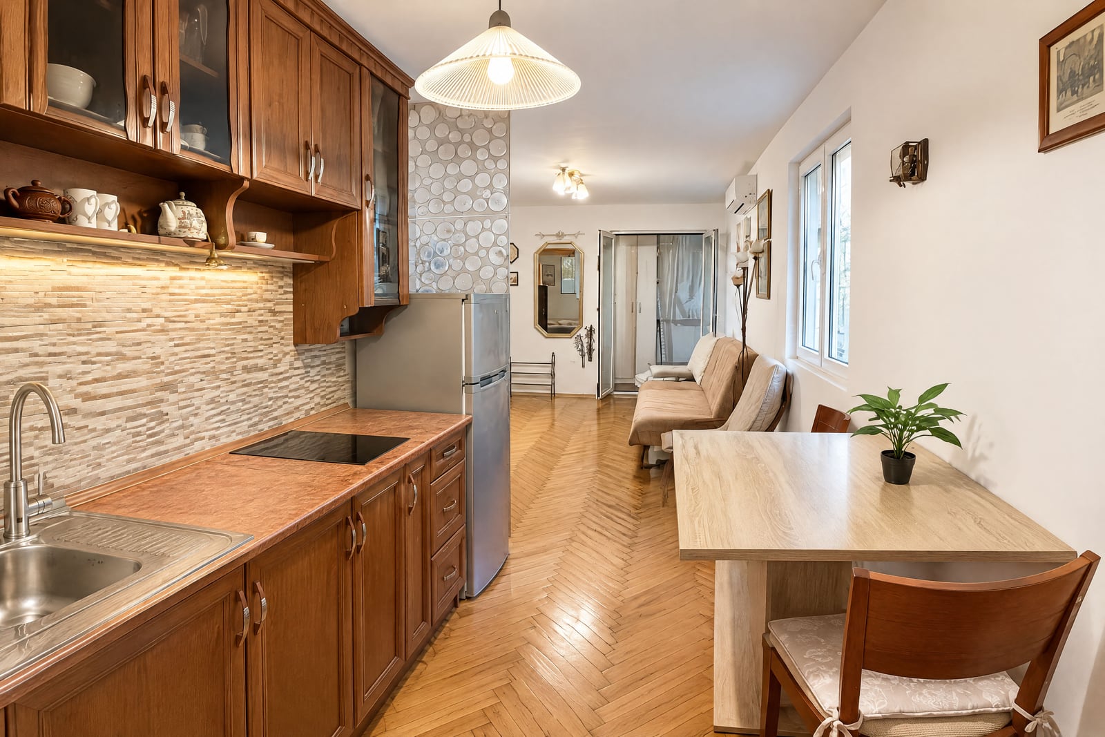 Kitchen with wooden cabinets, sink, stove, and fridge; dining table and sofa visible on herringbone parquet floor.