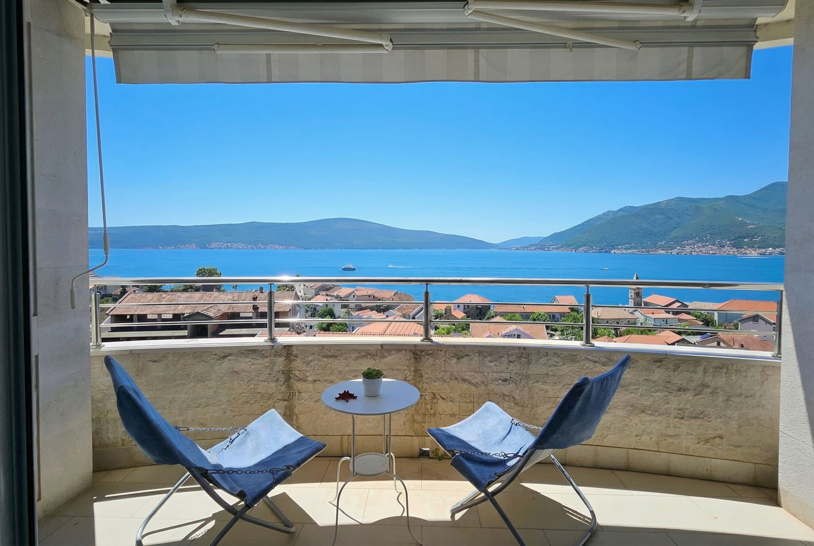 Balcony with panoramic sea and mountain views in an apartment, Tivat, Montenegro: two blue chairs and a table, stone finish.