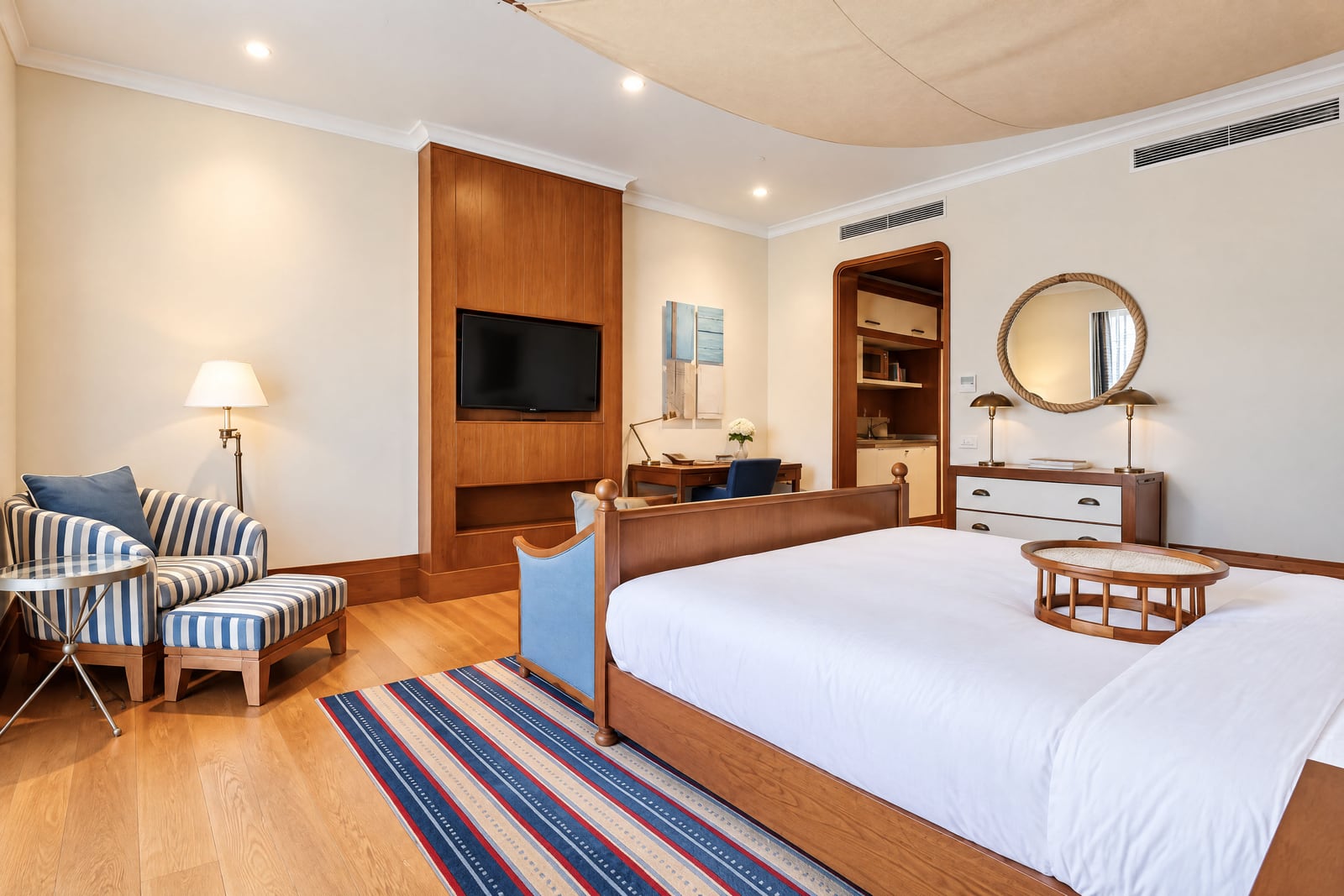 Bedroom featuring a wooden bed, striped armchair, TV in a wood niche, and a round rope mirror.