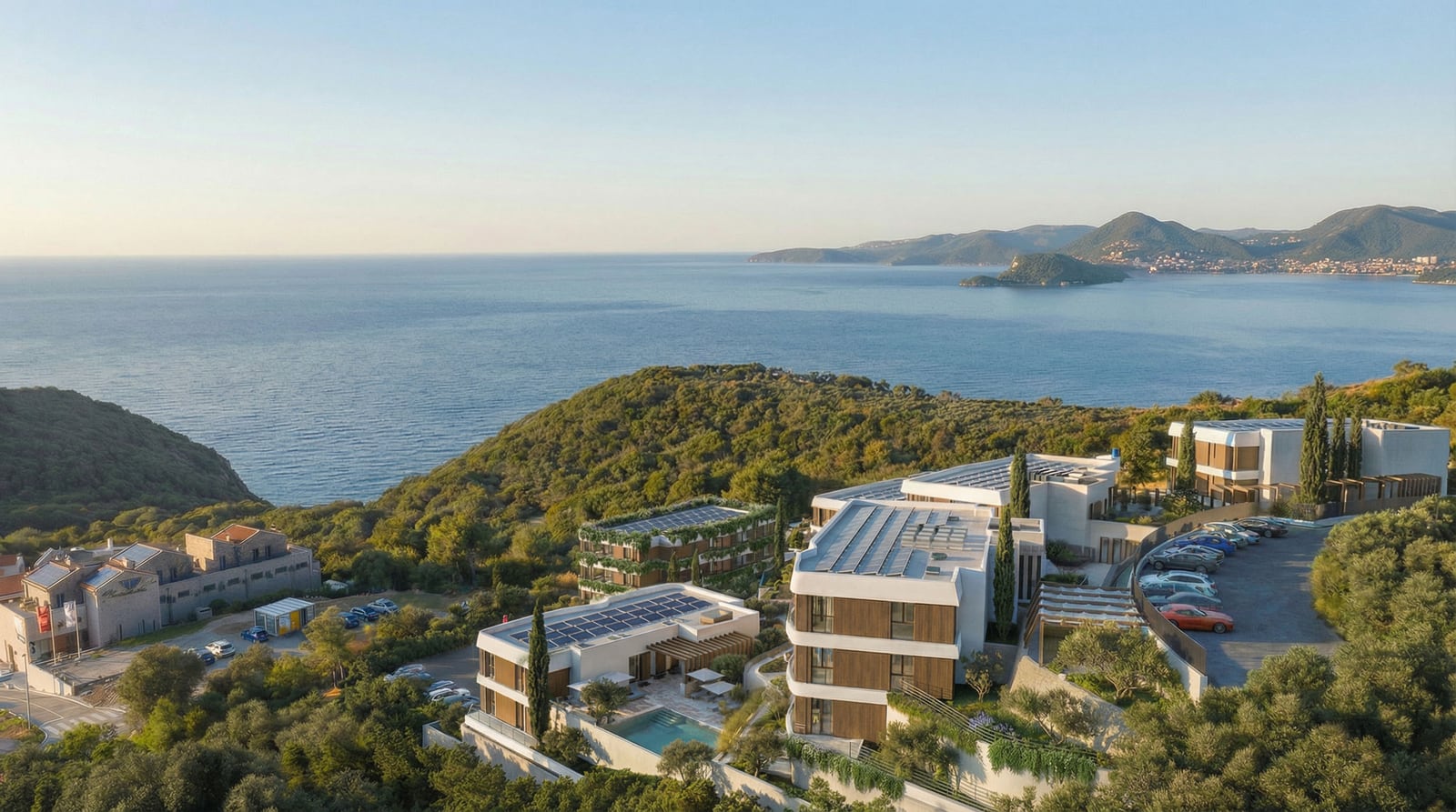 Aerial view of a modern coastal hotel complex with a pool, surrounded by trees and sea.