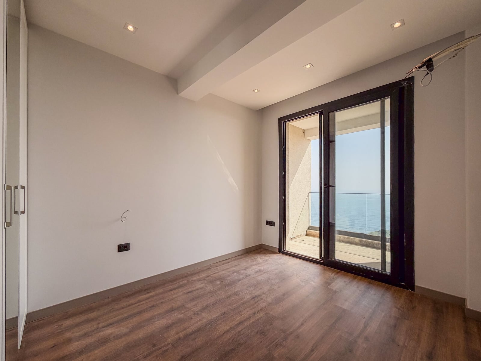 Empty room with wood flooring and white walls. A glass door leads to a balcony overlooking the sea.