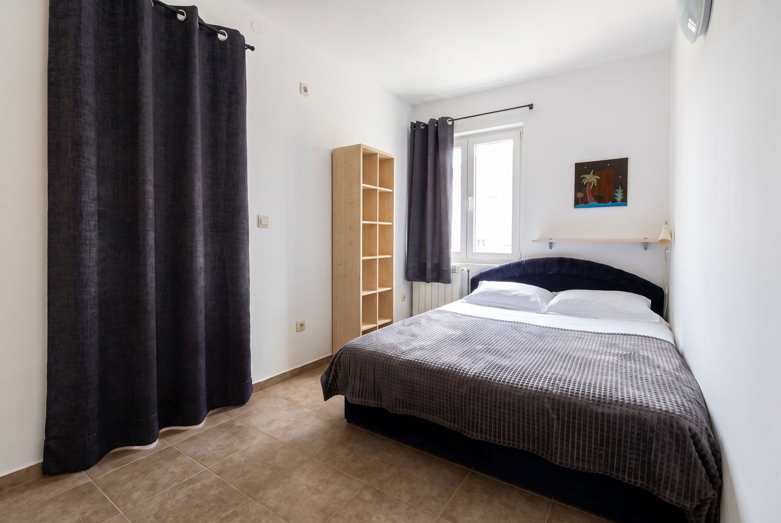 Bedroom featuring a double bed with grey cover, wooden shelving unit, dark curtains, and tiled flooring.