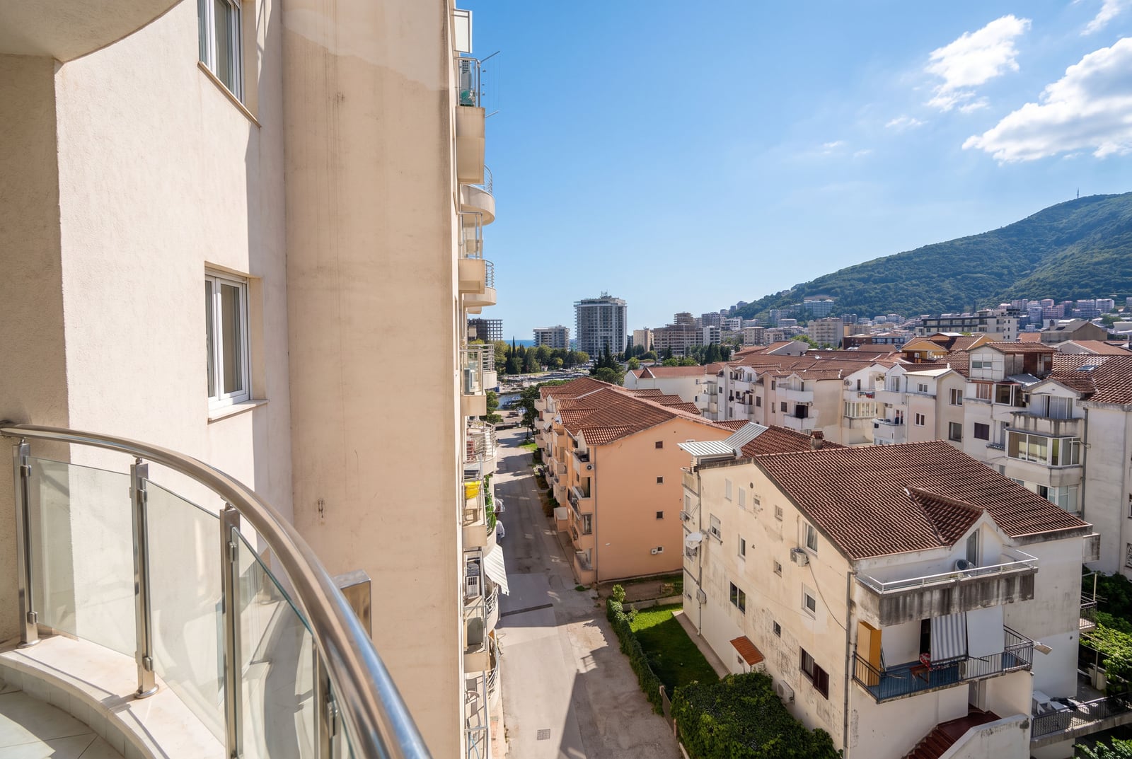 View from a balcony with glass railing overlooking a city street, residential buildings, and mountains.
